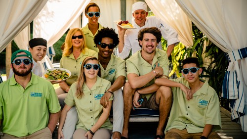 Nine students sitting under a cabana. Eight students are wearing bright green shirts and one student is wearing a chef's uniform.